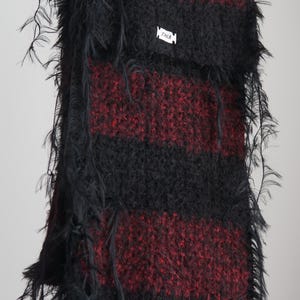 May include: A long, fuzzy scarf featuring alternating horizontal stripes of black and deep red. The knit fabric has a textured, shaggy appearance with loose, wispy fibres along the edges. A small white rectangular tag with the text ZAKA is attached.