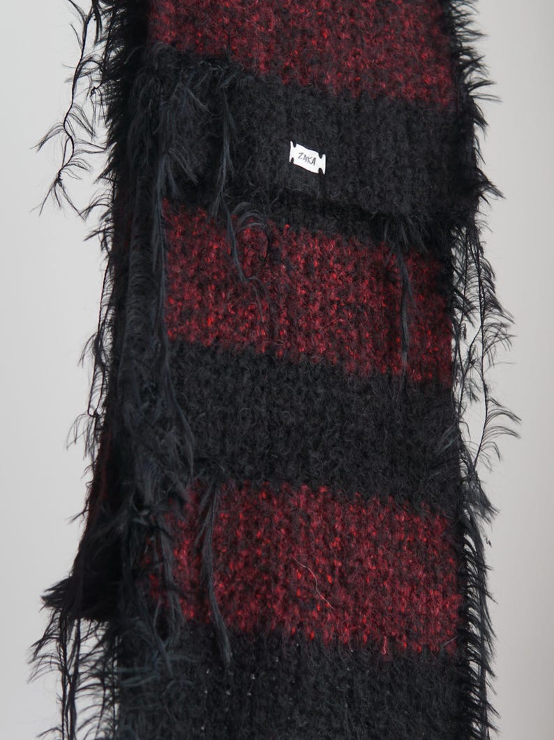 May include: A long, fuzzy scarf featuring alternating horizontal stripes of black and deep red. The knit fabric has a textured, shaggy appearance with loose, wispy fibres along the edges. A small white rectangular tag with the text ZAKA is attached.