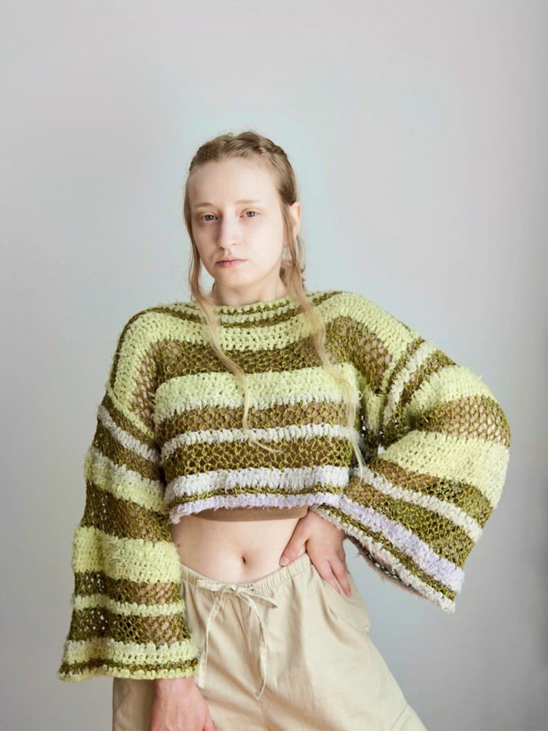 May include: A cropped, long-sleeved crochet jumper featuring horizontal stripes in shades of light green, olive green, and a hint of lavender. The garment has a loose, open-knit texture with wide, flared sleeves and a round neckline.