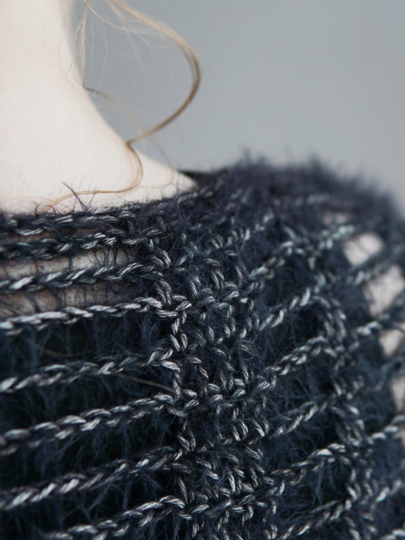 May include: A dark navy blue and silver metallic knit sweater featuring a loose, open-weave crochet pattern. The garment is crafted from a fuzzy, textured yarn that creates a soft, hairy appearance across the surface of the interlocking stitches.