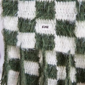 May include: A crochet scarf featuring a checkered pattern in dark forest green and off-white. The yarn has a fuzzy, textured appearance. A small white rectangular fabric label is sewn onto the center with the black text ZAIKA printed on it.