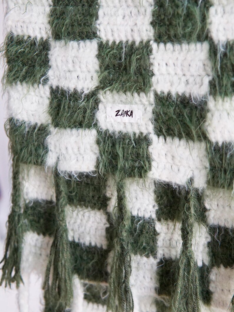 May include: A crochet scarf featuring a checkered pattern in dark forest green and off-white. The yarn has a fuzzy, textured appearance. A small white rectangular fabric label is sewn onto the center with the black text ZAIKA printed on it.