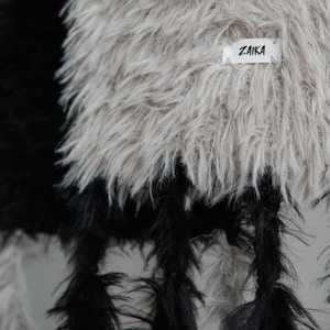 May include: A textured garment featuring long, shaggy faux fur in contrasting black and light beige sections. The beige section includes a small white rectangular fabric label with the black text ZAIKA stitched onto the surface of the material.
