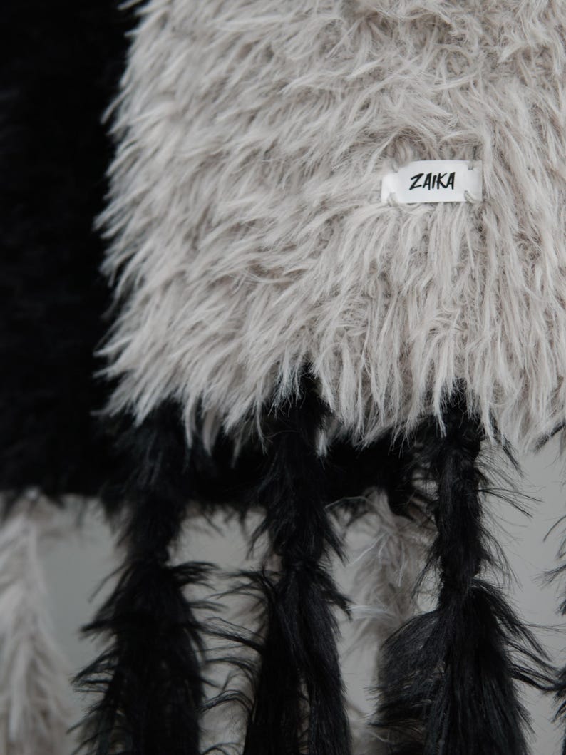 May include: A textured garment featuring long, shaggy faux fur in contrasting black and light beige sections. The beige section includes a small white rectangular fabric label with the black text ZAIKA stitched onto the surface of the material.