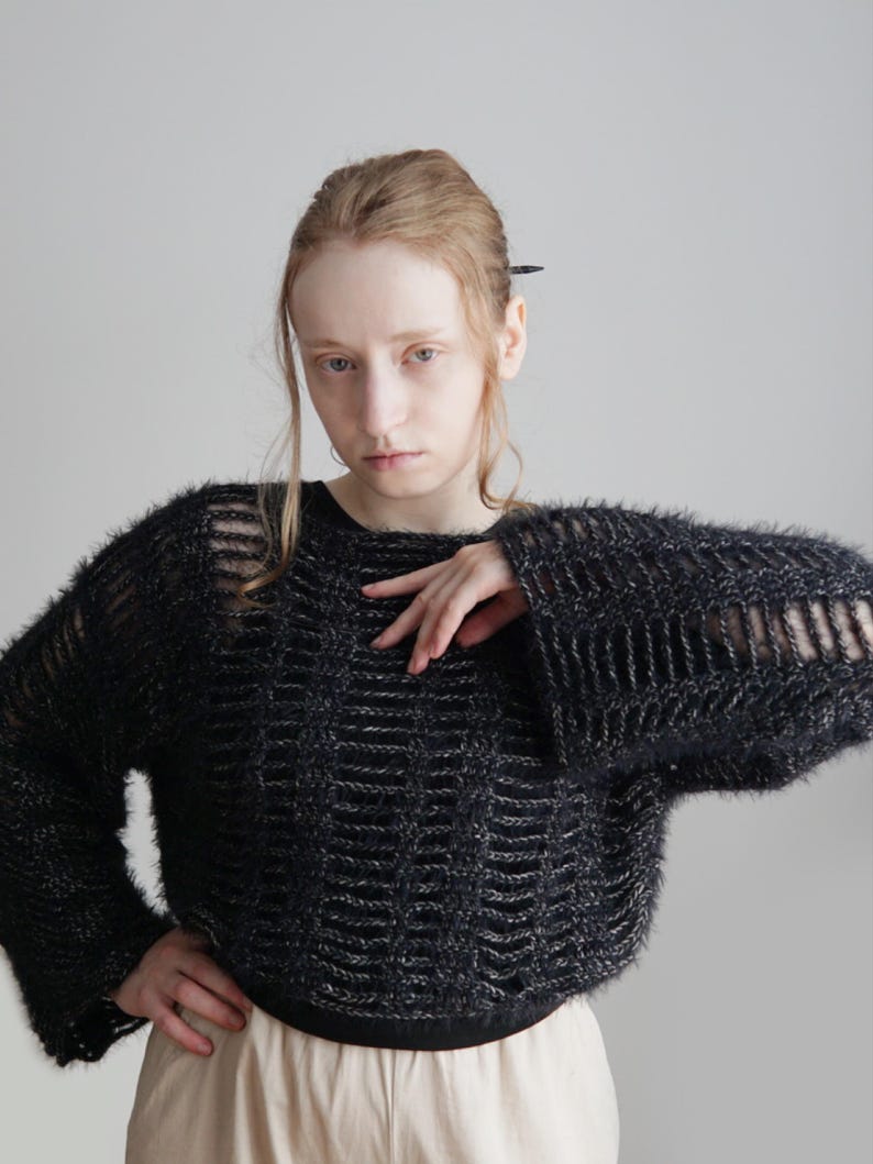 May include: A black fuzzy knit sweater featuring a wide-gauge, open-weave grid pattern throughout the body and sleeves. The garment has a round neckline, long sleeves with a flared silhouette, and a solid black hemline at the waist.