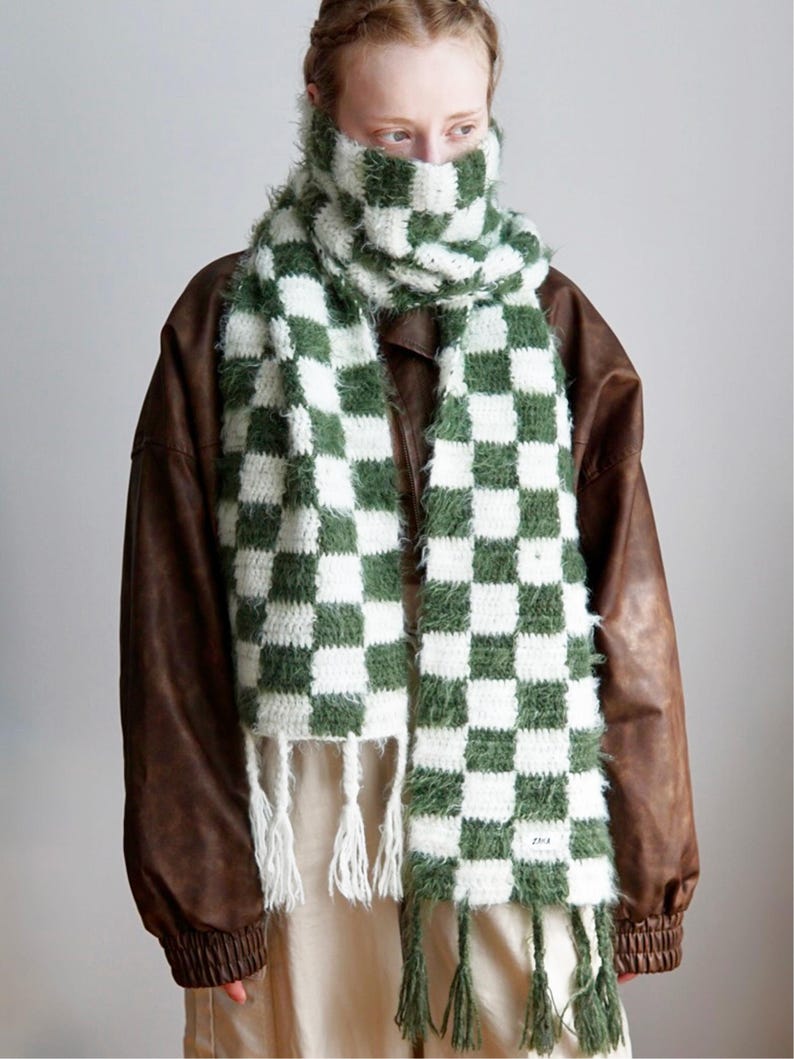 May include: A long, fuzzy, crocheted scarf featuring a checkered pattern in forest green and white. The scarf has a textured, yarn-like appearance with fringe detailing at both ends and a small rectangular white label on the lower right corner.