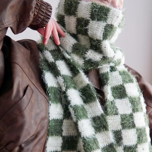 May include: A long, rectangular crochet scarf featuring a checkered pattern in alternating white and olive green squares. The scarf is constructed with a combination of smooth yarn and a fuzzy, textured yarn, creating a soft, tactile appearance.