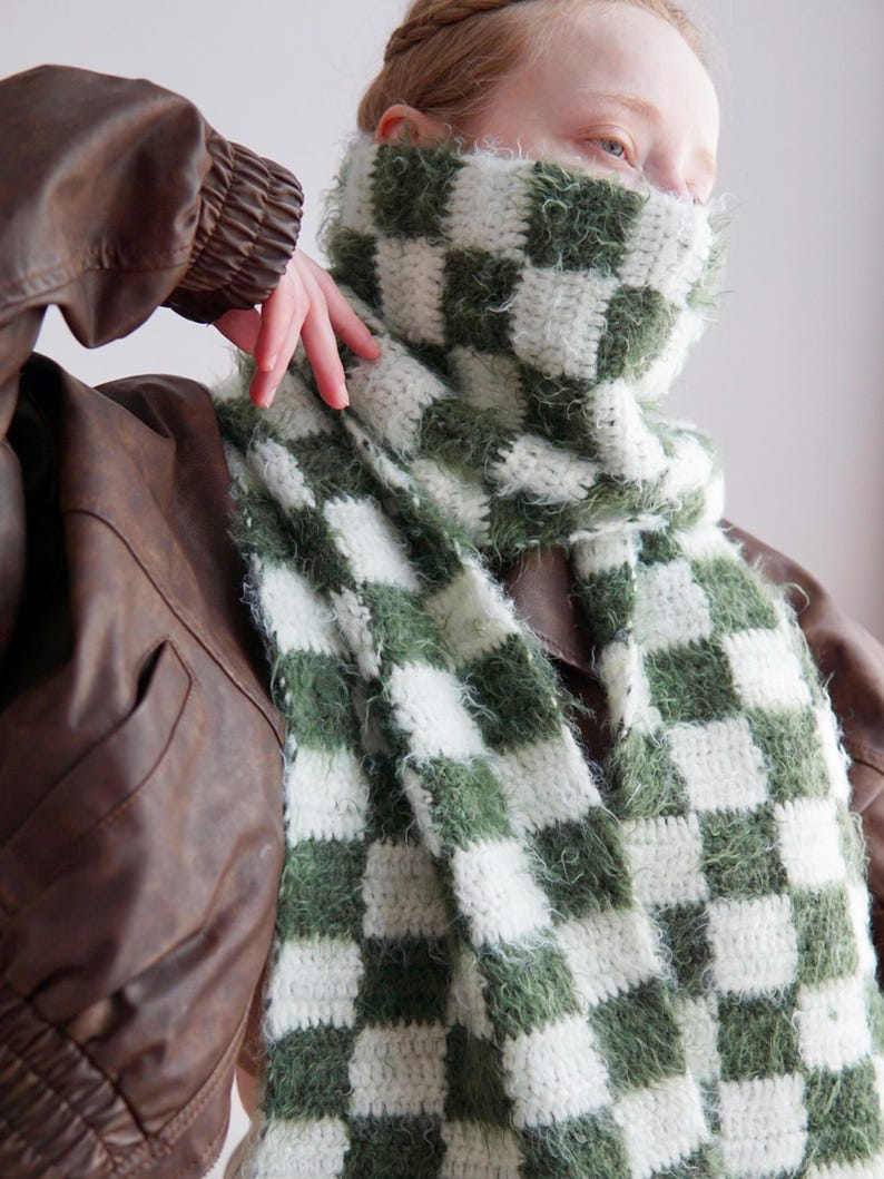 May include: A long, rectangular crochet scarf featuring a checkered pattern in alternating white and olive green squares. The scarf is constructed with a combination of smooth yarn and a fuzzy, textured yarn, creating a soft, tactile appearance.