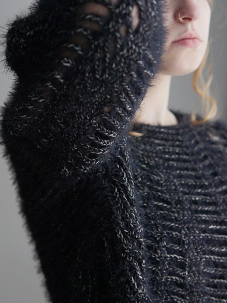 May include: A dark navy blue and silver fuzzy knit sweater featuring a textured, open-stitch pattern throughout the fabric. The garment has long sleeves and a round neckline, constructed from a soft, shaggy yarn with metallic silver accents.