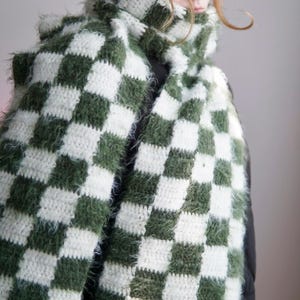 May include: A long, crocheted scarf featuring a repeating checkered pattern in forest green and white. The scarf is constructed from a combination of smooth yarn and fuzzy, textured yarn, creating a varied surface. A small white label is attached.