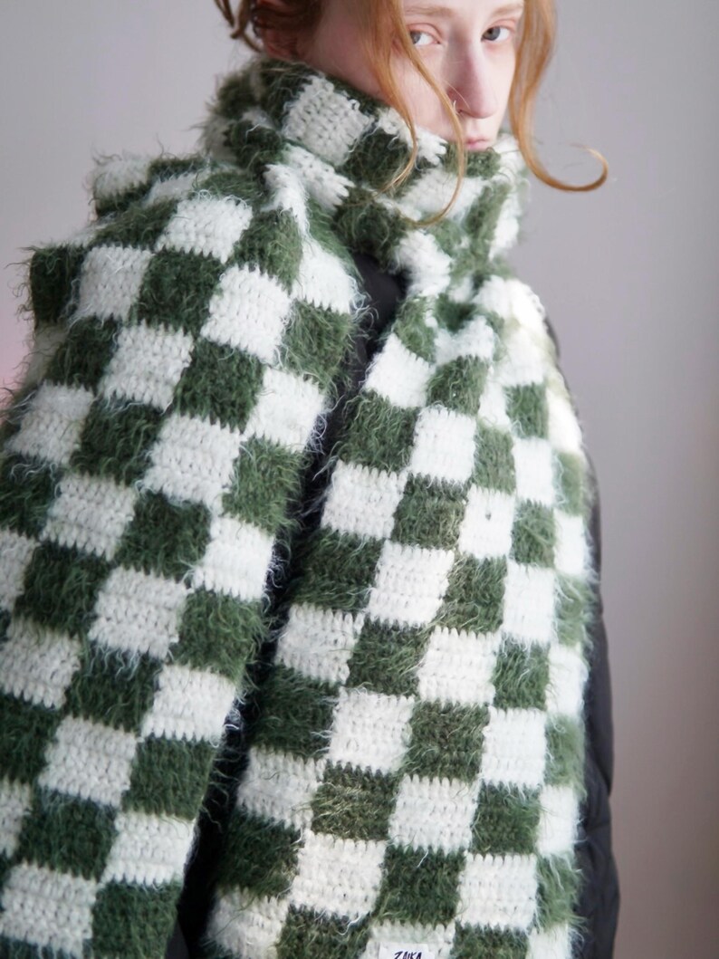 May include: A long, crocheted scarf featuring a repeating checkered pattern in forest green and white. The scarf is constructed from a combination of smooth yarn and fuzzy, textured yarn, creating a varied surface. A small white label is attached.