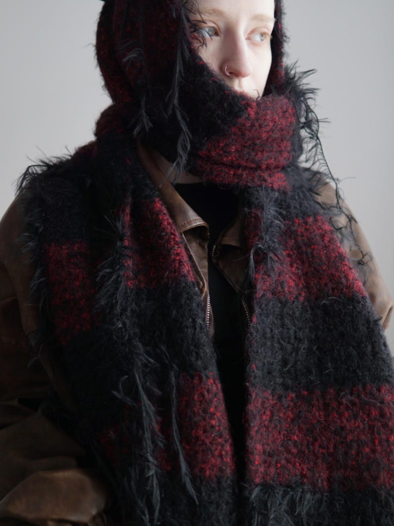 May include: A fuzzy, textured scarf featuring a bold, alternating plaid pattern in deep black and dark red. The scarf has a loose, shaggy knit construction with visible frayed edges and is wrapped around the head and neck over a brown jacket.