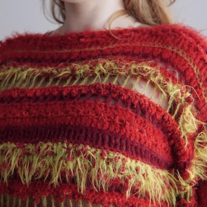 May include: A long-sleeve sweater featuring horizontal stripes in shades of red and chartreuse. The garment is constructed with a mix of dense, textured crochet stitches and open, airy sections, incorporating fuzzy, elongated yarn fibers throughout.