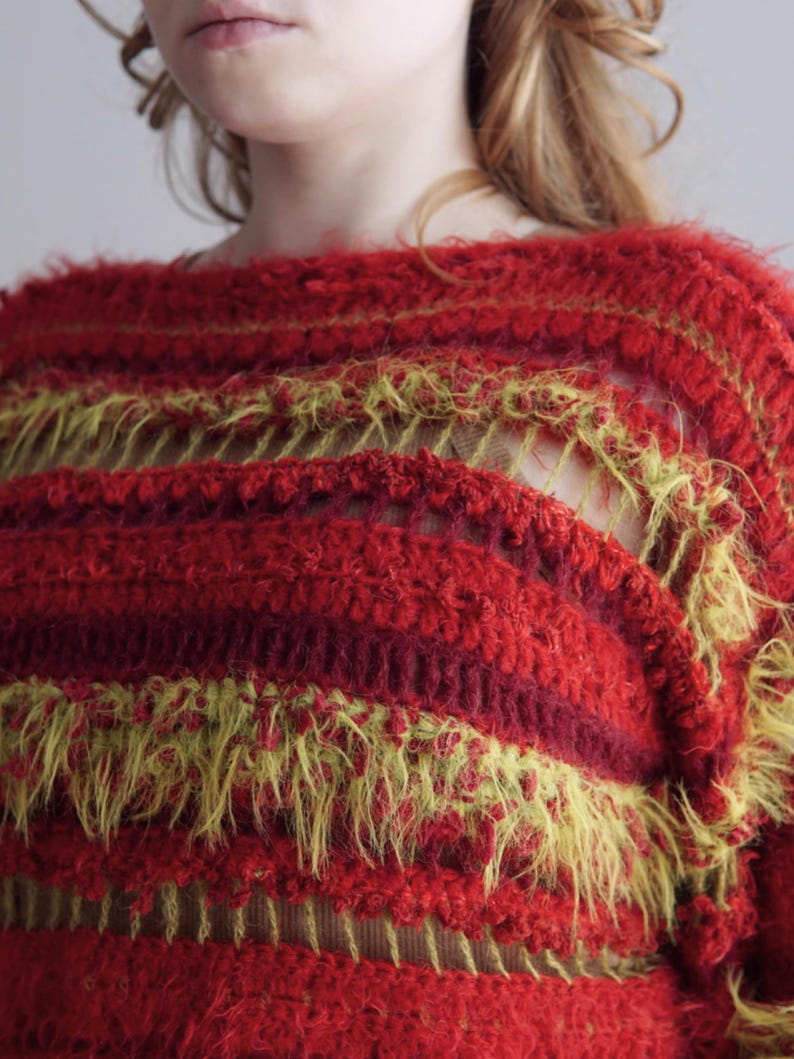 May include: A long-sleeve sweater featuring horizontal stripes in shades of red and chartreuse. The garment is constructed with a mix of dense, textured crochet stitches and open, airy sections, incorporating fuzzy, elongated yarn fibers throughout.