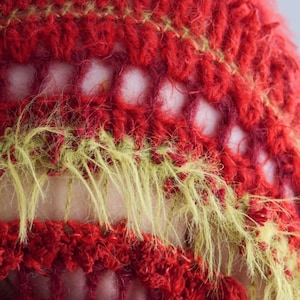 May include: A close-up of a crocheted textile featuring alternating horizontal bands of deep red yarn and fuzzy, textured chartreuse green yarn. The pattern includes open, airy crochet stitches that create a grid-like structure between the solid rows.