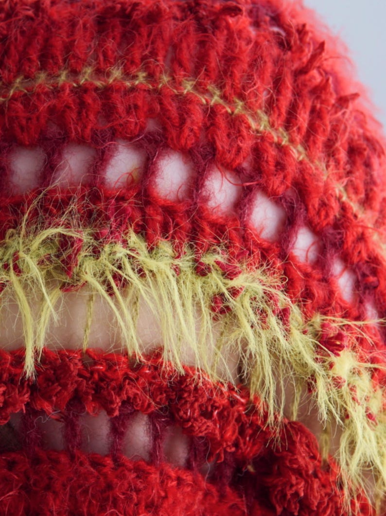 May include: A close-up of a crocheted textile featuring alternating horizontal bands of deep red yarn and fuzzy, textured chartreuse green yarn. The pattern includes open, airy crochet stitches that create a grid-like structure between the solid rows.