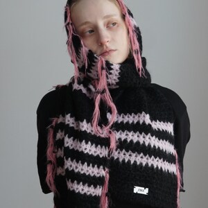 May include: A black and light pink striped knit hooded scarf featuring a fuzzy, textured yarn construction with loose, frayed pink fringe accents along the edges and a small white rectangular label with black text reading ZAKA.