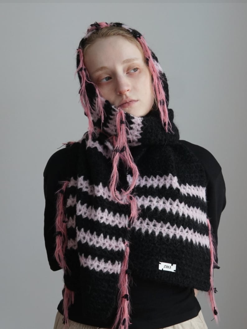May include: A black and light pink striped knit hooded scarf featuring a fuzzy, textured yarn construction with loose, frayed pink fringe accents along the edges and a small white rectangular label with black text reading ZAKA.
