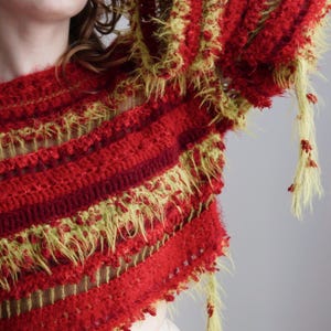 May include: A long-sleeved crochet sweater featuring alternating horizontal bands of deep red and fuzzy lime green yarn. The garment has a textured, open-knit construction with loose, shaggy yarn strands hanging from the sleeves and hemline.