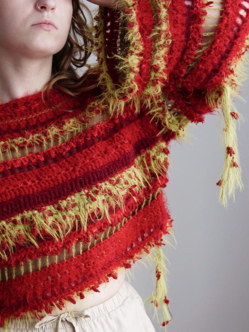 May include: A long-sleeved crochet sweater featuring alternating horizontal bands of deep red and fuzzy lime green yarn. The garment has a textured, open-knit construction with loose, shaggy yarn strands hanging from the sleeves and hemline.