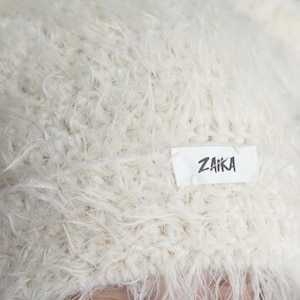 May include: A cream-colored beanie made from a fuzzy, shaggy yarn texture. The hat features a rectangular white fabric label sewn onto the front with the word ZAIKA printed in a bold, black, hand-drawn style font across the center.