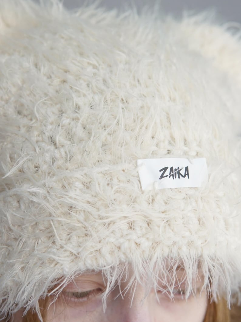 May include: A cream-colored beanie made from a fuzzy, shaggy yarn texture. The hat features a rectangular white fabric label sewn onto the front with the word ZAIKA printed in a bold, black, hand-drawn style font across the center.