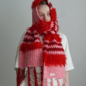 May include: A hooded scarf featuring a crochet knit pattern in shades of red and pink. The accessory includes textured, fuzzy yarn sections, long fringe detailing at the ends, and a small white rectangular label with black text on the lower edge.