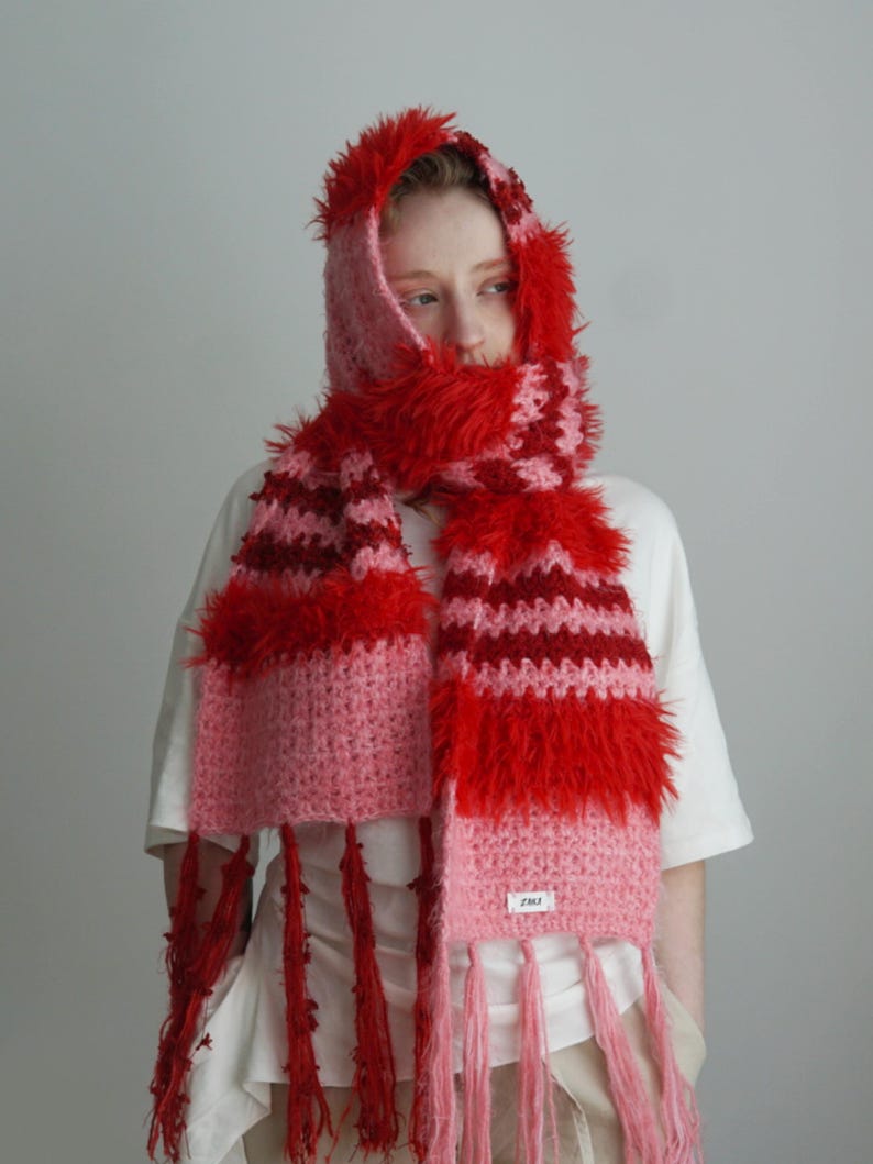 May include: A hooded scarf featuring a crochet knit pattern in shades of red and pink. The accessory includes textured, fuzzy yarn sections, long fringe detailing at the ends, and a small white rectangular label with black text on the lower edge.