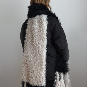 May include: A long, rectangular scarf featuring a color-blocked design with a wide central panel of shaggy, light beige faux fur flanked by sections of shaggy black faux fur. The ends of the scarf are finished with long, dangling fringe tassels.