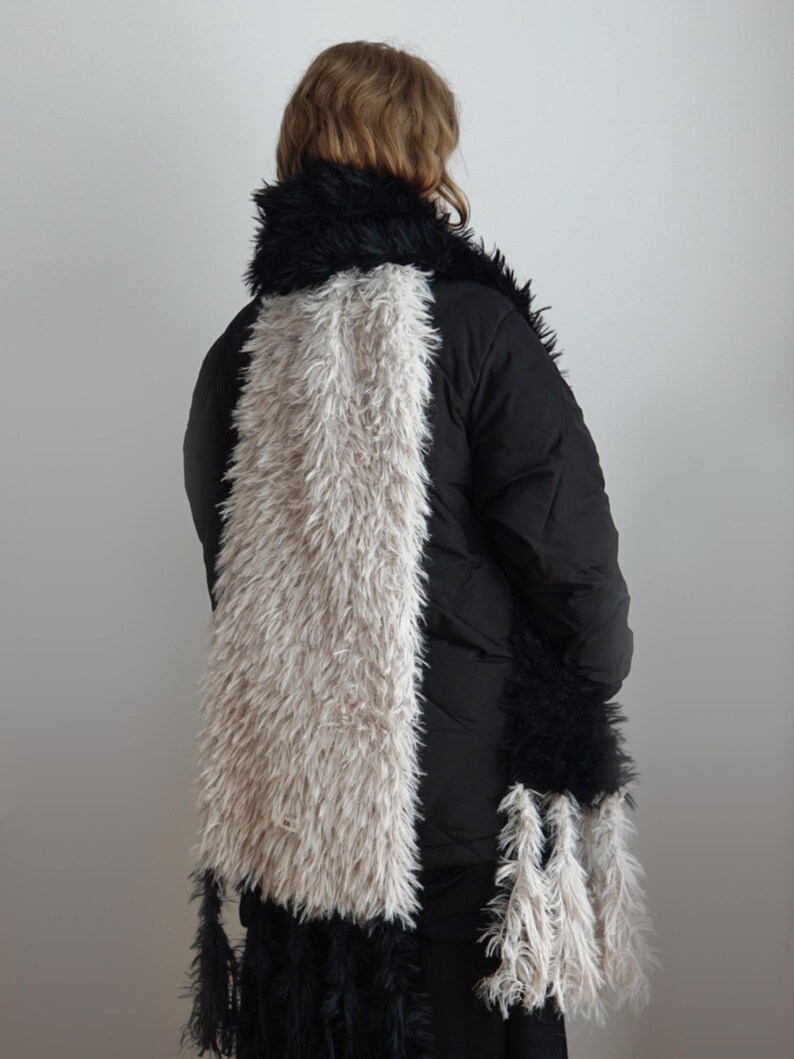 May include: A long, rectangular scarf featuring a color-blocked design with a wide central panel of shaggy, light beige faux fur flanked by sections of shaggy black faux fur. The ends of the scarf are finished with long, dangling fringe tassels.