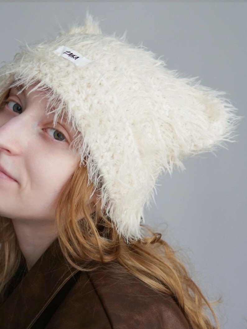 May include: A cream-colored, fuzzy, textured beanie hat with a pointed cat-ear shape. The hat features a small white rectangular fabric label on the front with the black text ZAIKA printed on it. The material has a long, soft, shaggy pile.