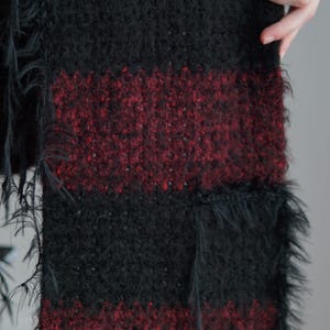 May include: A knitted scarf featuring alternating horizontal stripes of solid black and a deep burgundy red. The texture is fuzzy and slightly uneven, with loose, frayed black yarn fibres extending from the edges of the fabric throughout the length.