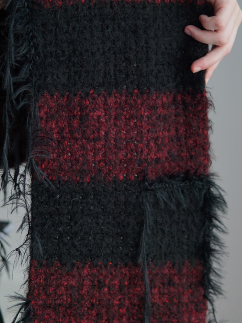 May include: A knitted scarf featuring alternating horizontal stripes of solid black and a deep burgundy red. The texture is fuzzy and slightly uneven, with loose, frayed black yarn fibres extending from the edges of the fabric throughout the length.