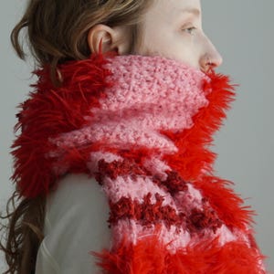 May include: A chunky knit scarf featuring alternating horizontal stripes of soft light pink yarn and vibrant red fuzzy, textured yarn. The scarf is wrapped around the neck, showcasing a mix of smooth crochet stitches and long, shaggy fibres.