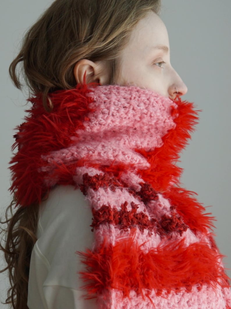 May include: A chunky knit scarf featuring alternating horizontal stripes of soft light pink yarn and vibrant red fuzzy, textured yarn. The scarf is wrapped around the neck, showcasing a mix of smooth crochet stitches and long, shaggy fibres.