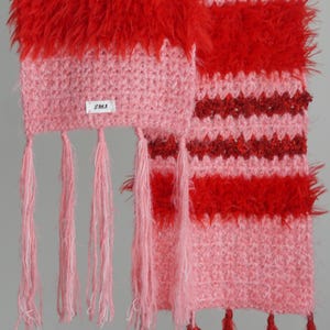 May include: A crochet scarf featuring alternating horizontal stripes of light pink and vibrant red yarn. The texture varies between smooth stitches and fuzzy, shaggy sections. A small white label with the text ZAIKA is attached. Pink and red tassels hang from the ends.
