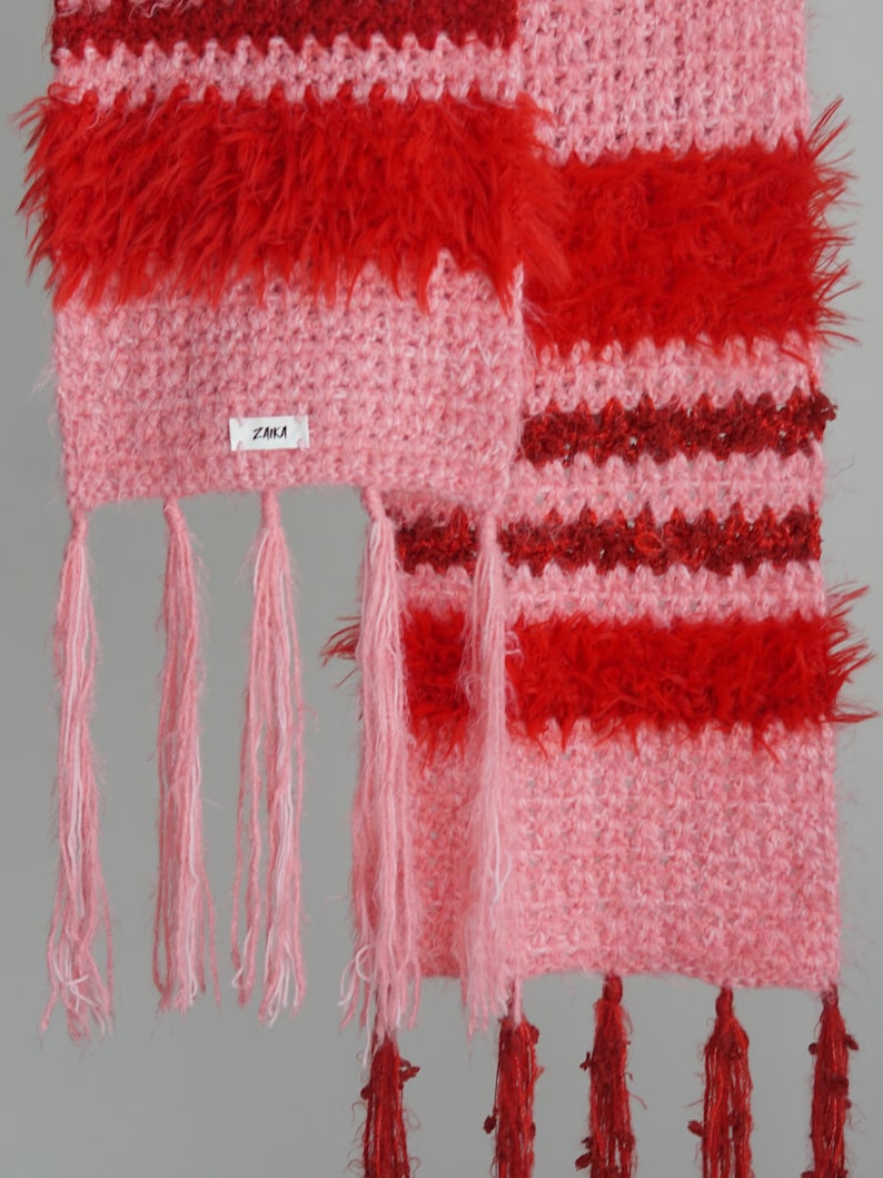May include: A crochet scarf featuring alternating horizontal stripes of light pink and vibrant red yarn. The texture varies between smooth stitches and fuzzy, shaggy sections. A small white label with the text ZAIKA is attached. Pink and red tassels hang from the ends.