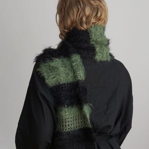 May include: A fuzzy, long scarf featuring alternating horizontal stripes of black and olive green. The scarf has a textured, mohair-like appearance with visible knit loops and a soft, hairy surface, draped around the neck over a solid black shirt.