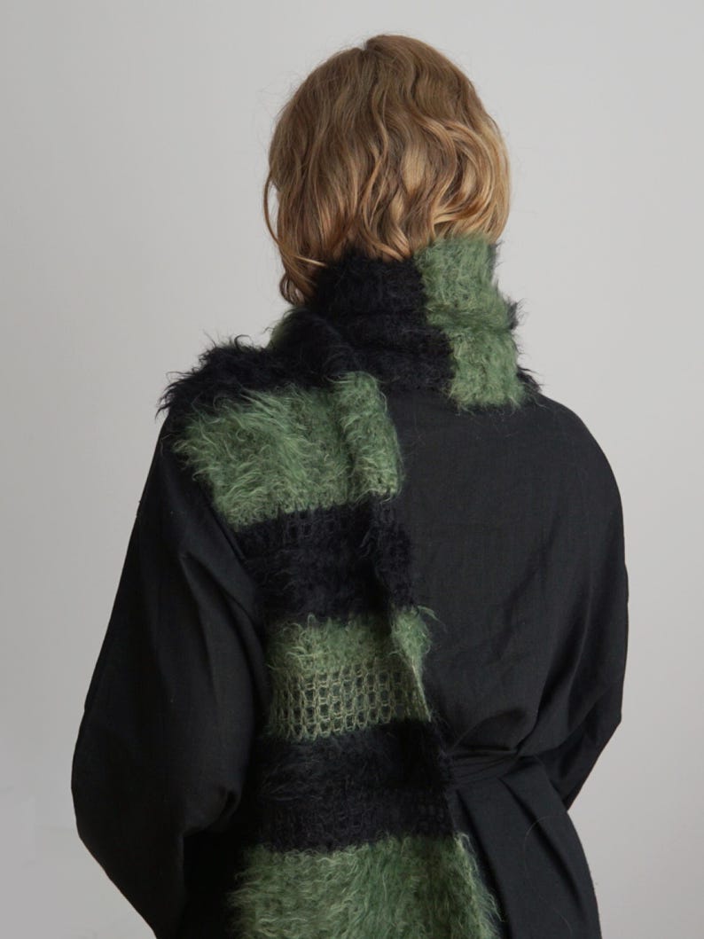 May include: A fuzzy, long scarf featuring alternating horizontal stripes of black and olive green. The scarf has a textured, mohair-like appearance with visible knit loops and a soft, hairy surface, draped around the neck over a solid black shirt.