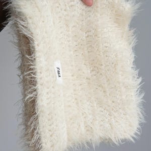 May include: A cream-colored, shaggy, faux-fur infinity scarf with a long-pile texture. The scarf features a small, rectangular white fabric label sewn onto the front with the black text Z.AIKA printed clearly on the surface of the tag.
