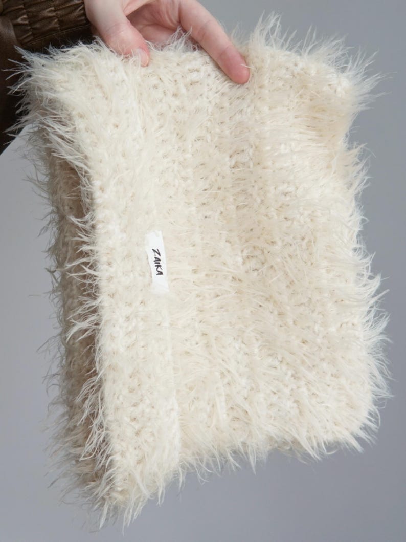 May include: A cream-colored, shaggy, faux-fur infinity scarf with a long-pile texture. The scarf features a small, rectangular white fabric label sewn onto the front with the black text Z.AIKA printed clearly on the surface of the tag.