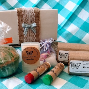 May include: A gift box filled with bath and body products. The box is wrapped in brown burlap and white lace. The products include a bath bomb, a lotion bar, a lip balm, and a bar of soap. The soap is labeled "Wood Rose" and is from "Eastern Organics Ltd Naturals".