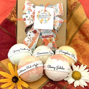 May include: A fall-themed gift box with a decorative bow. The box contains several bath bombs labeled "Granny Smith Apple," "Cinnamon Spice," "Pumpkin Pie," "Autumn Red Apple," and "Cherry Jubilee." A yellow flower and a white daisy are also present.