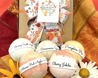 May include: A fall-themed gift box with a decorative bow. The box contains several bath bombs labeled "Granny Smith Apple," "Cinnamon Spice," "Pumpkin Pie," "Autumn Red Apple," and "Cherry Jubilee." A yellow flower and a white daisy are also present.