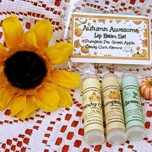 May include: An autumn-themed lip balm set with three flavors: Candy Corn, Pumpkin Pie, and Green Apple. The set includes lip balm tubes, a small pumpkin, and candy corn decorations. A large yellow sunflower is also present.