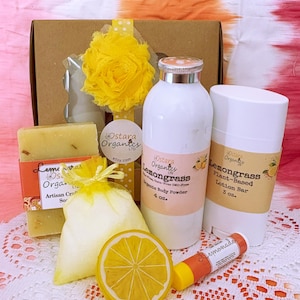 May include: A gift box filled with lemongrass-scented bath and body products. The box includes a bar of soap, a bag of bath salts, a bottle of body powder, a deodorant stick, and a lip balm. The products are all packaged in white containers with yellow and green labels. The box is decorated with a yellow bow and a yellow flower.