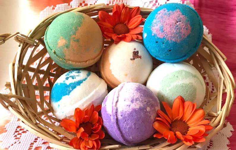 May include: A wicker basket filled with six colorful bath bombs, each with a unique swirl pattern. The bath bombs are surrounded by orange flowers.
