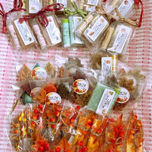 May include: A collection of individually wrapped gift sets containing soaps and lip balms. Some sets feature leaf-shaped soaps, while others include rectangular bars and tubes tied with colorful ribbons, twine, and artificial autumn berries.
