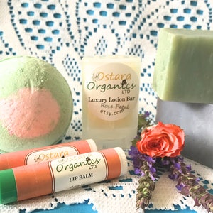 May include: A bath bomb, two lip balms, a lotion bar, and two bars of soap. The bath bomb is green and pink. The lip balms are green and orange. The lotion bar is white with the text "Ostara Organics LTD Luxury Lotion Bar Rose Petal etsy.com". The soap bars are green and gray.