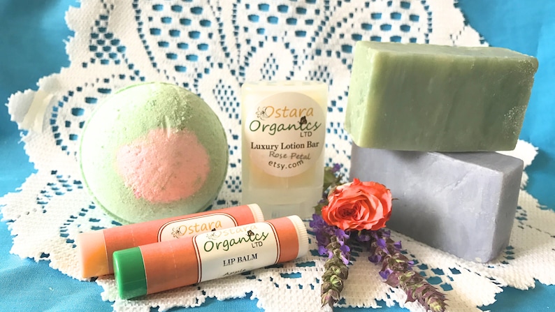 May include: A bath bomb, two lip balms, a lotion bar, and two bars of soap. The bath bomb is green and pink. The lip balms are green and orange. The lotion bar is white with the text "Ostara Organics LTD Luxury Lotion Bar Rose Petal etsy.com". The soap bars are green and gray.